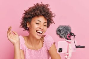 positive-pretty-brunette-afro-american-female-blogger-focused-smartphone-tripod-makes-online-streaming-has-own-channel-smiles-gladfully-poses-against-pink-wall-vlogging-concept